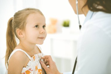 Bir çocuğu steteskopla muayene eden bir doktor. Parlak renkli elbise giymiş mutlu gülümseyen kız hasta her zamanki sağlık kontrolünde.