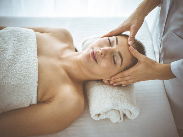 Beautiful brunette woman enjoying facial massage with closed eyes comfortable and blissful. Relaxing treatment in medicine and spa center concepts