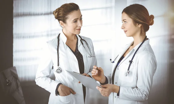 Two female physicians are discussing medical therapy, while standing at the table in a sunny clinic office. Doctors use pc computer at work. Teamwork in medicine