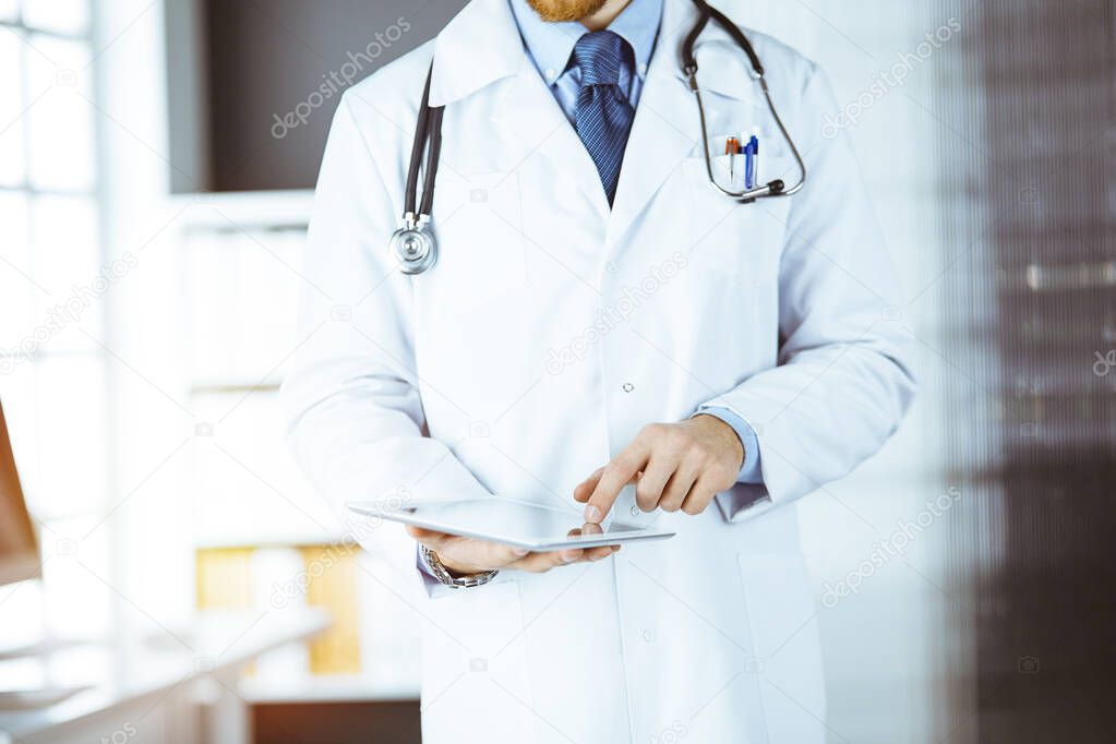 Friendly bearded doctor using tablet computer in sunny clinic at his working place