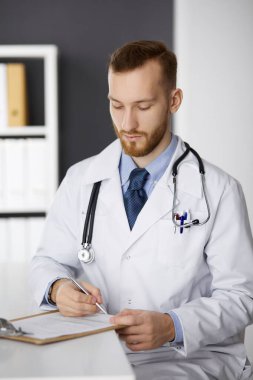 Dost canlısı kızıl sakallı doktor klinikteki panoda oturuyor ve yazıyor. Hastanede mükemmel bir tıbbi hizmet. İlaç, sağlık hizmetleri