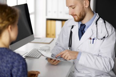 Dost canlısı kızıl sakallı doktor ve hasta kadın klinikte oturup tablet bilgisayar kullanırken sağlık muayenesini tartışıyorlar.