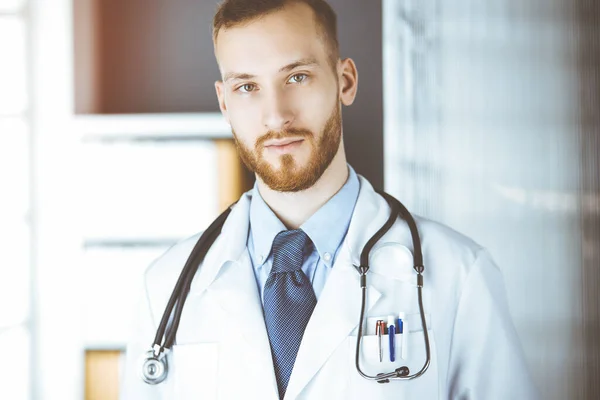 Κοκκινογένης γιατρός στέκεται ευθεία στην ηλιόλουστη κλινική κοντά στο χώρο εργασίας του. Έννοια ιατρικής — Φωτογραφία Αρχείου