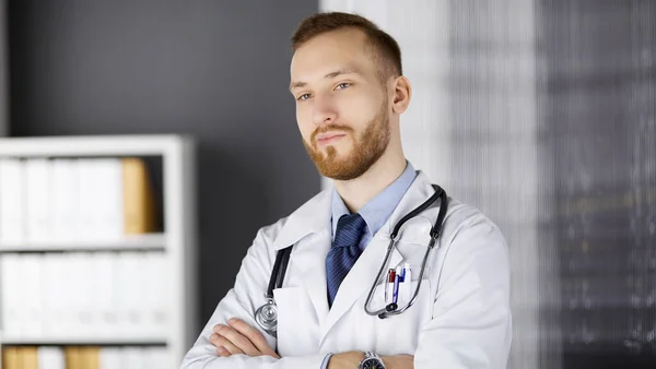 Kızıl sakallı doktor çalışma yerinin yanındaki klinikte dikiliyor. Hekim portresi. İlaç, sağlık hizmetleri — Stok fotoğraf