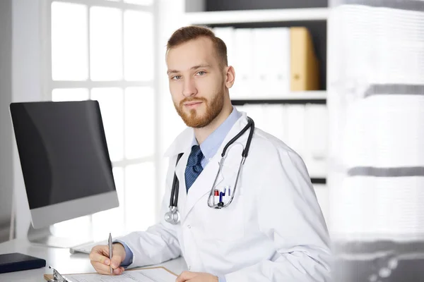 Amigável médico de barba vermelha sentado e escrevendo na área de transferência na clínica. Serviço médico perfeito no hospital. Medicina, cuidados de saúde — Fotografia de Stock