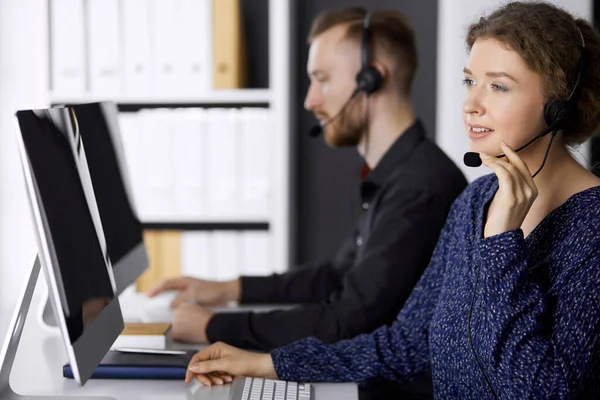 Femme d'affaires parlant par casque tout en étant assis avec un collègue à barbe rouge dans le bureau moderne. Télémarketing et service à la clientèle. Groupe de personnes diversifiées dans le centre d'appels — Photo