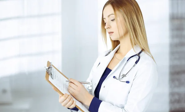 Jovem médica está fazendo algumas anotações usando uma prancheta, enquanto está em seu gabinete em uma clínica. Retrato de bela médica com um estetoscópio. Serviço médico perfeito em um hospital — Fotografia de Stock