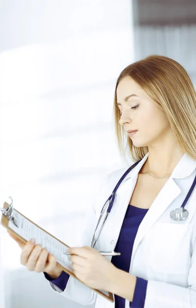 Молода розумна жінка-лікар призначає пацієнту ліки, використовуючи буфер обміну, стоячи в кабінеті в клініці. Жінка-лікар зі стетоскопом на робочому місці — стокове фото