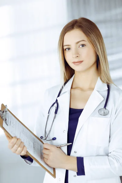 Giovane donna-medico intelligente sta prescrivendo alcune medicine per un paziente, utilizzando un blocco appunti, mentre in piedi nell'armadio in una clinica. Medico femminile con stetoscopio sul posto di lavoro — Foto Stock