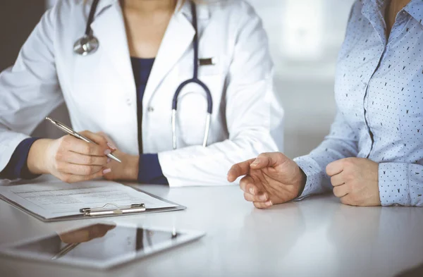 Mulher desconhecida- médico está ouvindo seu paciente, enquanto se sentam juntos na mesa no armário em uma clínica. Médica com um estetoscópio está escrevendo na área de transferência, de perto. Perfeito médico — Fotografia de Stock