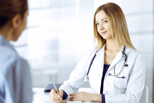 Jeune femme-médecin intelligente écoute les symptômes de ses patients, tout en étant assis ensemble dans le cabinet dans une clinique. Médecin, avec un sourire, écrit quelques notes sur ses papiers à la — Photo