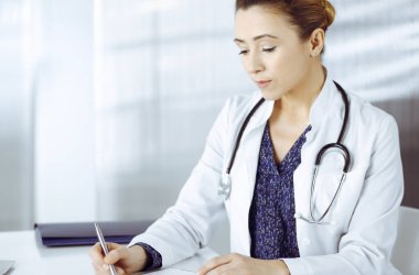 Neşeli, zeki bir kadın doktor klinikteki dolabının masasında otururken bir pano kullanıyor ve bazı ilaç isimleri yazıyor. Profesyonel kadın doktor portresi.