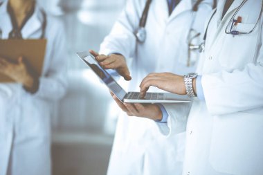 Steteskoplu bir grup tanınmamış doktor, erkek ve kadın hastane ofisinde dikilirken dizüstü bilgisayar kullanarak muayene sonuçlarını tartışıyor. Doktorlar ilaçları kontrol etmek için dizüstü bilgisayar kullanıyorlar.