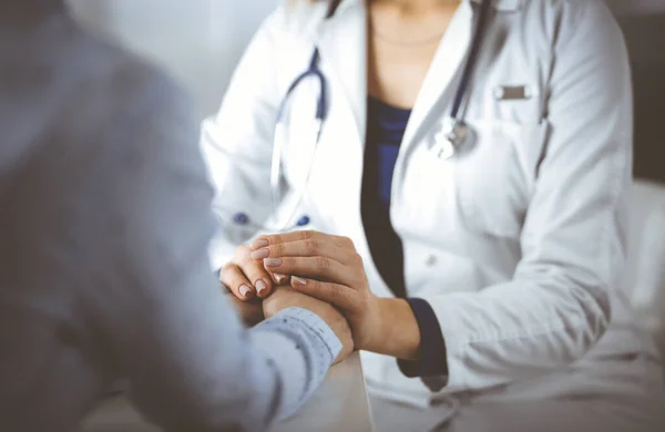 Unknown woman-doctor is holding her patients hands to reassure the patient, discussing current health examination, while they are sitting together at the desk in the cabinet in a clinic. Female — Stock Photo, Image