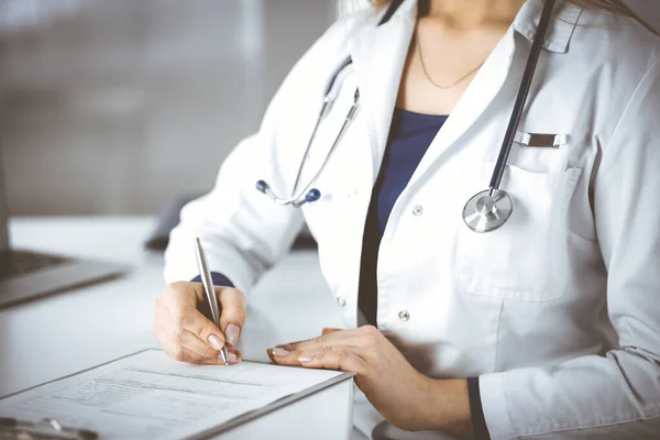 Une femme-médecin inconnue prescrit des médicaments à sa patiente, à l'aide d'un presse-papiers, assise au bureau de son cabinet. Médecine avec stéthoscope, gros plan. Service médical parfait — Photo