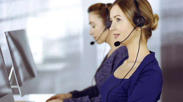 Two busineswomen have conversations with the clients by headsets, while sitting at the desk in a modern office. Diverse people group in a call center. Telemarketing and customer service — Stock Photo, Image