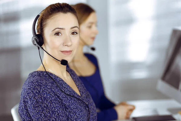 Two busineswomen have conversations with the clients by headsets, while sitting at the desk in a modern office. Diverse people group in a call center. Telemarketing and customer service