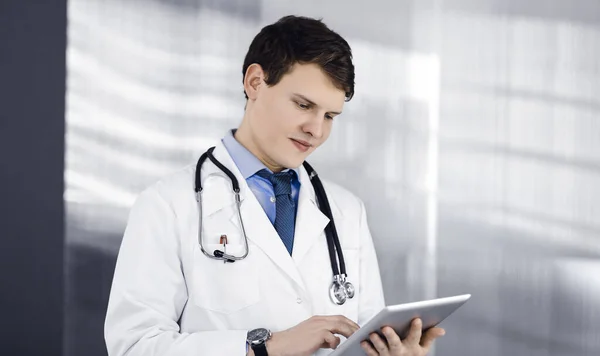 Mladý přátelský doktor kontroluje nějaké informace na svém počítači. Portrét profesionálního lékaře v práci na klinice. Koncept medicíny — Stock fotografie