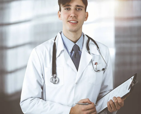 Přátelský mladý lékař, stojící s podložkou na moderní klinice. Koncept medicíny — Stock fotografie