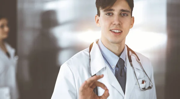 Přátelský muž-doktor stojí zpříma a ukazuje na klinice ceduli OK. Kolegyně má naspěch na pozadí lékaře. Koncept medicíny — Stock fotografie