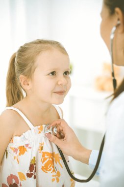Bir çocuğu steteskopla muayene eden bir doktor. Parlak renkli elbise giymiş mutlu gülümseyen kız hasta her zamanki sağlık kontrolünde.