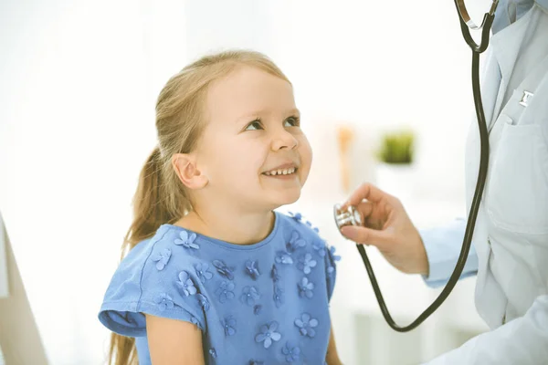 Doctor examinando a un niño por estetoscopio. Feliz niña sonriente paciente vestida con vestido azul es en la inspección médica habitual. Concepto de medicina —  Fotos de Stock