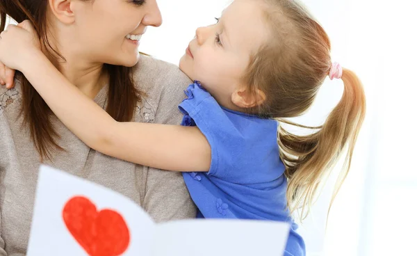 Concept de fête des mères. Enfant fille félicite maman et donne sa carte postale en forme de coeur rouge. Maman et fille heureux souriant et étreignant. Plaisir et vacances en famille — Photo