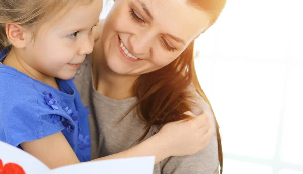 Muttertagskonzept. Die Tochter gratuliert der Mutter und überreicht ihr eine Postkarte mit rotem Herzchen. Mutter und Mädchen glücklich lächelnd und umarmt. Familienspaß und Urlaub — Stockfoto