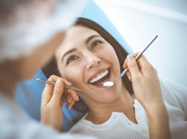 Gülümseyen esmer kadın diş hekimi tarafından muayene ediliyor. Hastanın ağzının yanında diş aletleri tutan bir doktorun elleri. Sağlıklı diş ve ilaç konsepti