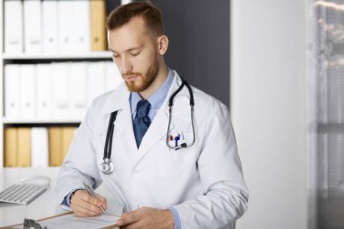 Dost canlısı kızıl sakallı doktor klinikteki panoda oturuyor ve yazıyor. Hastanede mükemmel bir tıbbi hizmet. İlaç, sağlık hizmetleri