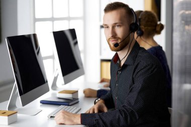 Modern ofiste otururken kulaklıkla konuşan sakallı iş adamı. Çağrı merkezinde çeşitli insanlar var. Tele pazarlama ve müşteri hizmetleri