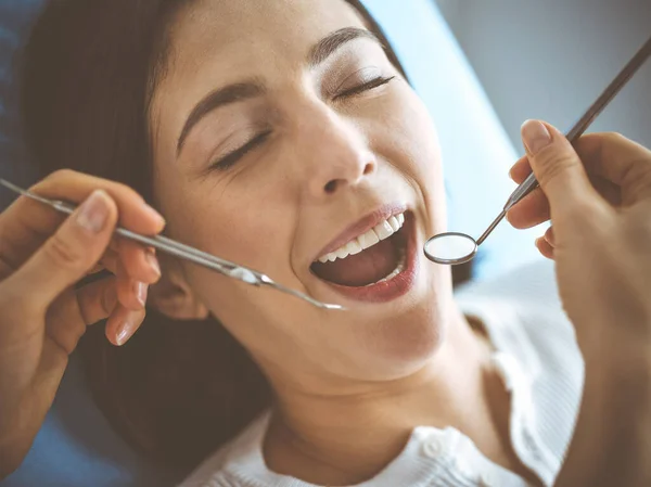 Smiling brunette woman being examined by dentist at dental clinic. Hands of a doctor holding dental instruments near patients mouth. Healthy teeth and medicine concept — Stock Photo, Image
