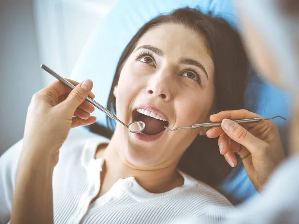 Uśmiechnięta brunetka badana przez dentystę w klinice dentystycznej. Ręce lekarza trzymającego instrumenty stomatologiczne w pobliżu ust pacjenta. Zdrowe zęby i koncepcja medycyny — Zdjęcie stockowe