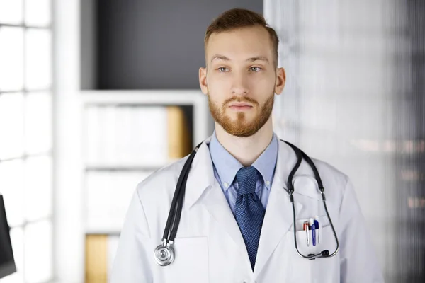 Κοκκινογένης γιατρός στέκεται ακριβώς στην κλινική κοντά στο χώρο εργασίας του. Πορτρέτο γιατρού. Ιατρική, υγειονομική περίθαλψη — Φωτογραφία Αρχείου