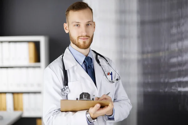 Φιλικός γιατρός με κόκκινα γένια στέκεται και γράφει με ντοσιέ στην κλινική στο χώρο εργασίας του. Τέλεια ιατρική υπηρεσία στο νοσοκομείο. Ιατρική, υγειονομική περίθαλψη — Φωτογραφία Αρχείου