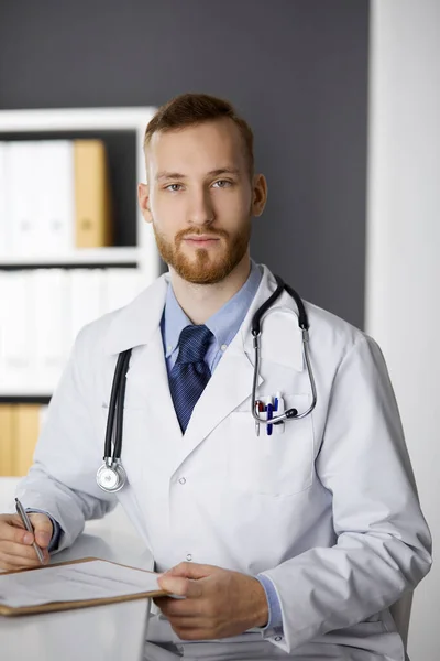 Amigável médico de barba vermelha sentado e escrevendo na área de transferência na clínica. Serviço médico perfeito no hospital. Medicina, cuidados de saúde — Fotografia de Stock