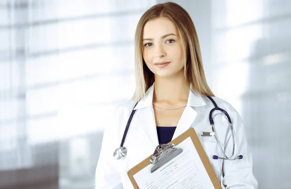 Giovane donna-medico ottimista tiene in mano una cartellina, mentre si trova in una clinica. Ritratto di medico donna amichevole con stetoscopio. Un servizio medico perfetto in ospedale. Medicina — Foto Stock