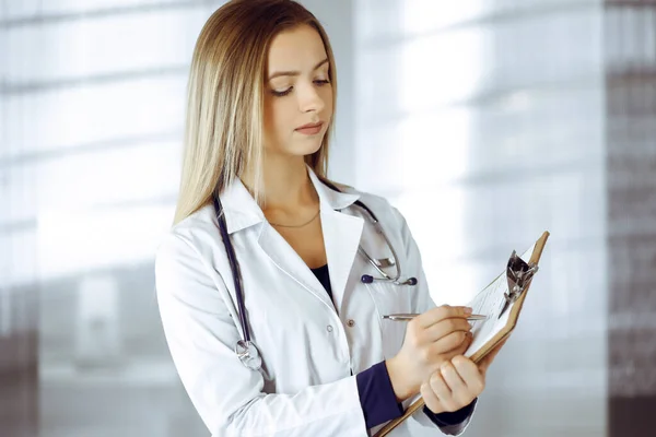 診療所のキャビネットに立っている間、若い女性医師はクリップボードを使用していくつかのメモを取っています。聴診器で美しい女性医師の肖像画。病院での完璧な医療サービス — ストック写真