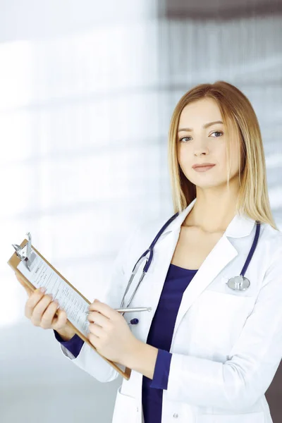 Una joven doctora inteligente está recetando algún medicamento para un paciente, usando un portapapeles, mientras está de pie en el gabinete de una clínica. Médico femenino con estetoscopio en su lugar de trabajo —  Fotos de Stock