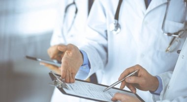 Steteskoplu bir grup doktor, erkek ve kadın hastane ofisinde ayakta dururken bir dosya ve tablet kullanarak tıbbi muayene sonuçlarını tartışıyor. Doktorlar ilaçları kontrol ediyor.