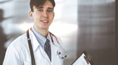 Modern klinikte dostane genç bir erkek doktor elinde bir panoyla dikiliyor. Bayan meslektaşım, doktorların arka planında ilaç kayıtları doldurmakla meşgul. Tıp konsepti