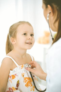 Bir çocuğu steteskopla muayene eden bir doktor. Parlak renkli elbise giymiş mutlu gülümseyen kız hasta her zamanki sağlık kontrolünde.