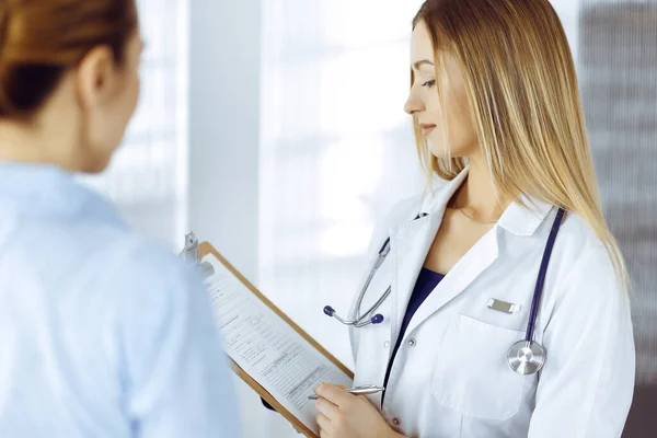 Jovem médica e sua paciente estão discutindo os pacientes exame de saúde atual, enquanto estão juntos em um consultório hospitalar. A médica está a escrever algumas marcas, usando uma prancheta. Perfeito. — Fotografia de Stock