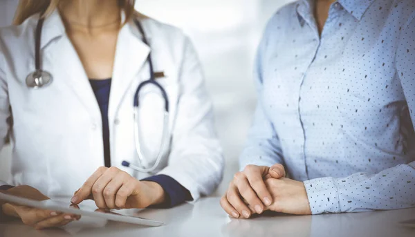 Mulher-médico desconhecida está mostrando a seu paciente uma descrição da medicação, enquanto se sentam juntos na mesa no armário em uma clínica. Médica feminina está usando um tablet de computador e um estetoscópio — Fotografia de Stock