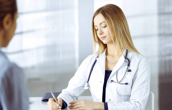 Junge intelligente Ärztin hört ihren Patienten zu, während sie in einer Klinik zusammen im Schrank sitzen. Ärztin mit Stethoskop schreibt an — Stockfoto