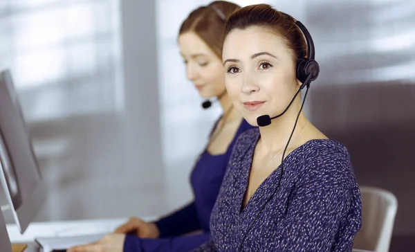 Dos mujeres tienen conversaciones con los clientes por auriculares, mientras están sentadas en el escritorio de una oficina moderna. Diversas personas se agrupan en un centro de llamadas. Telemarketing y servicio al cliente — Foto de Stock