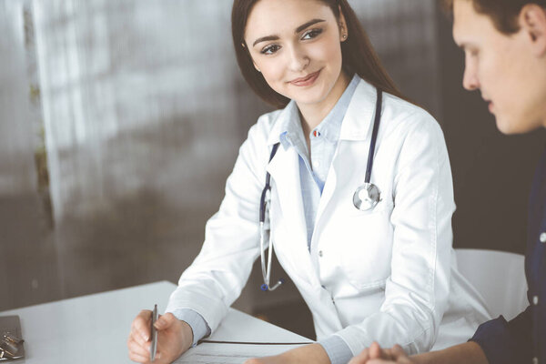 Friendly young woman-doctor is listening to her patient, while they are sitting together at the desk in a cabinet. Physician is holding a clipboard in her hands for filling up medication