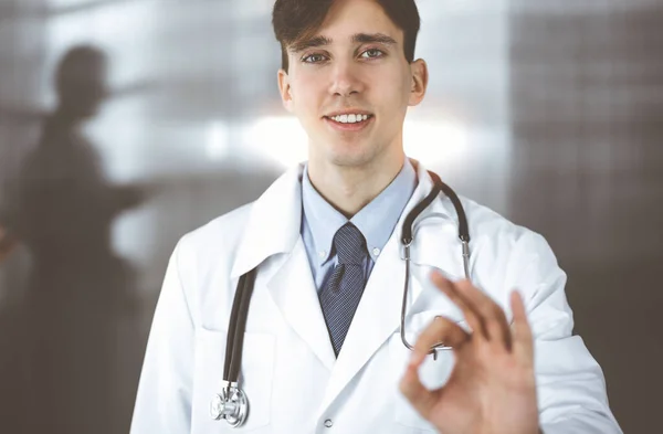 Amigável homem-médico de pé em linha reta e mostrando Ok sinal na clínica. Mulher colega está com pressa no fundo do médico. Conceito de medicina — Fotografia de Stock