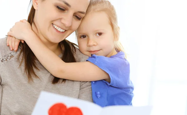 Концепція Дня матері. Дитяча дочка вітає маму і дає листівку з червоною формою серця. Мама і дівчинка щасливі посміхаються і обіймаються. Родинні веселощі та відпочинок — стокове фото