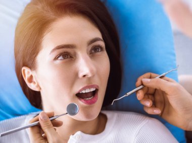 Güneşli diş kliniğinde dişçi tarafından muayene edilen gülümseyen esmer kadın. Hastanın ağzının yanında diş aletleri tutan bir doktorun elleri. Sağlıklı diş ve ilaç konsepti